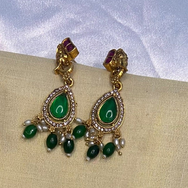 Premium gold-toned Lakshmi pendant necklace with elephant motifs, emerald green teardrop pendant, pearl accents, sparkling white stones, and matching earrings displayed on a cream jewelry bust. Traditional South Indian temple necklace perfect for weddings, Onam, Vishu, and festive occasions